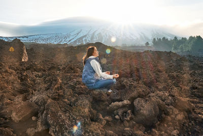 Woman enjoying freedom while meditating on lava stone at panoramic view of snowy summits of etna