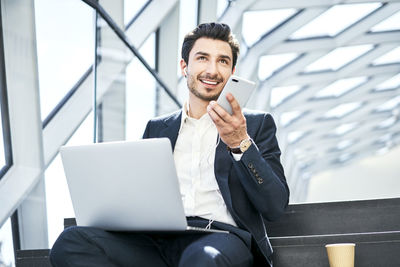 Smiling businessman sitting on stairs wearing earphones using cell phone and laptop