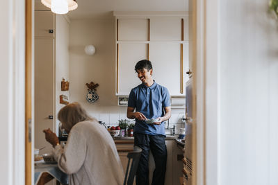 Smiling male caregiver talking to senior woman eating food in kitchen at home