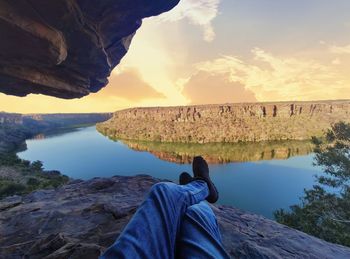 Low section of person on rock by lake against sky during sunset