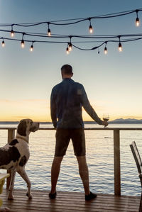 Man with dog standing against sea at sunset