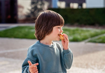 Hungry toddler boy enjoying snack while walking outside in park at spring sunny day.
