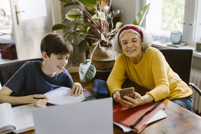 Smiling senior woman using smart phone while sitting with grandson doing homework at home