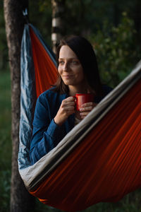 Female traveler is sitting in a hammock and drinking tea from a red cup. hiking outdoor recreation