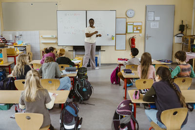 Happy female teacher standing against whiteboard with students in classroom