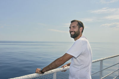 Young man standing on railing by sea against sky