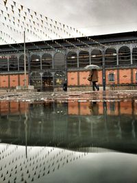 People walking with umbrellas against building reflecting in puddle