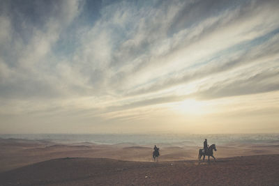 People riding horses on hill against cloudy sky during sunset