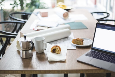 High angle view of teapot with documents rolled up by laptop and croissant on table