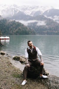 A tall brutal man hipster groom in a wedding suit stands by the misty mountains and lake in nature