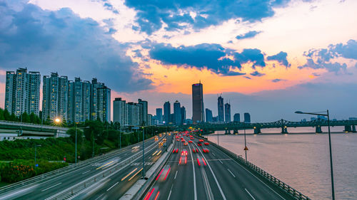 Light trails on road by river against sky during sunset