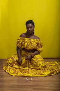 Young beautiful woman sitting on wooden floor, wearing colorful dress. against yellow background.