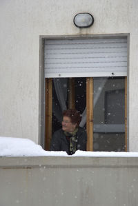 Woman looking away while standing by window during snowfall