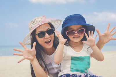 Happy mother and daughter wearing sunglasses enjoying at beach against sky