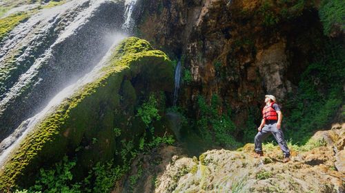 Man looking at waterfall while standing on rocks during sunny day