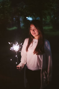 Young woman holding illuminated sparkler while standing at night