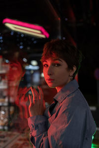 Sad lonely woman in red neon light leaning on shop window on street at night, looking at camera