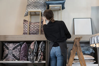 Rear view of young female entrepreneur standing on ladder while arranging cushion in rack at store