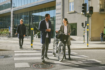 Full length of smiling man and woman talking while walking with electric bicycle on crosswalk against building in city