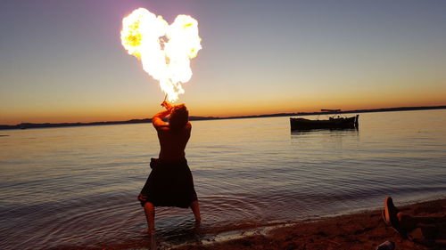 Rear view of fire-eater blowing fire while standing on shore at beach against sky