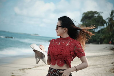 Young woman carrying high heels on beach against sky