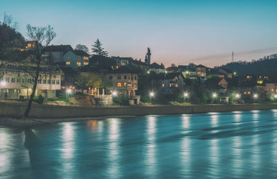 Illuminated buildings by river against sky at dusk