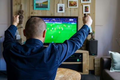 Rear view of man enjoying soccer match on television set in living room at home