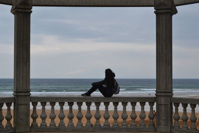 Side view of woman sitting on railing at beach against sky