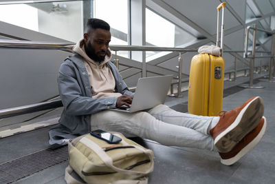 Intent young black guy with computer on laps sitting on airports floor booking apartment for journey