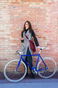 Smiling young woman with bike leaning on wall while using mobile