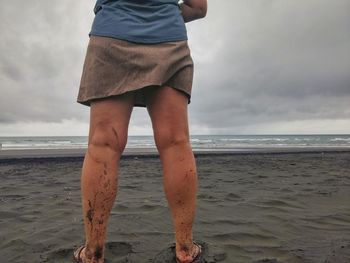 Low section of woman with dirty legs standing at beach against cloudy sky