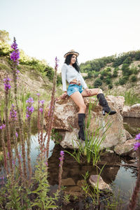 Young woman on rock by lake , plants against sky