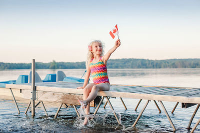 Full length of girl holding canadian flag sitting on pier by lake