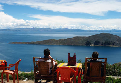 Rear view of man sitting on chair by lake against sky