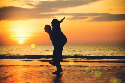 Silhouette man piggybacking woman while standing at beach against sky during sunset