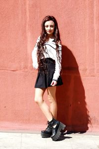 Full length portrait of young woman standing against wall on sunny day