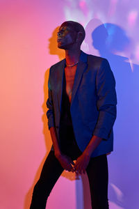 Confident bald african american slim male model with necklace in trendy outfit standing at wall with glowing lights in modern studio looking away