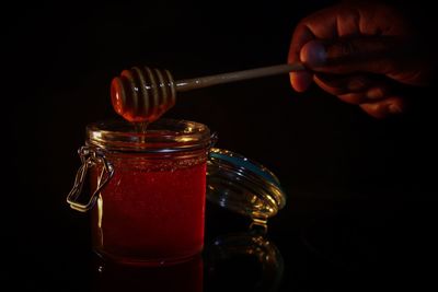 Cropped hand holding dipper over honey in jar against black background