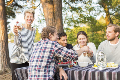 Portrait of happy woman holding beer glass while standing by friends having lunch at picnic table