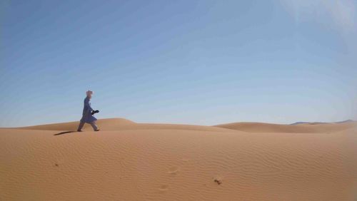 Side view of man in traditional clothing walking on sand dunes against blue sky