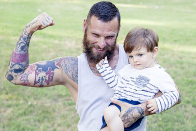 Smiling bearded man flexing muscle while holding son on field 