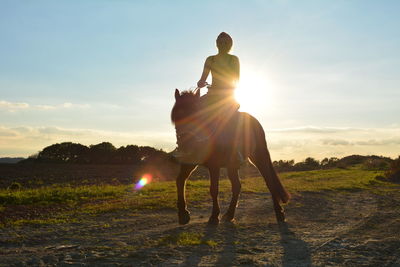 Woman riding horse on grassy field against sky during sunny day