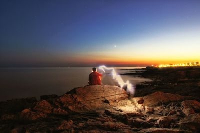 Rear view of man sitting on rock at beach against sky during sunset