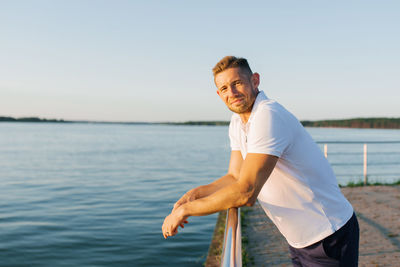 A smiling man in a white polo shirt leaning on a railing by a serene lake under soft evening light.