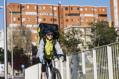 Cheerful female courier with thermal bag riding bike on bridge while delivering food in city