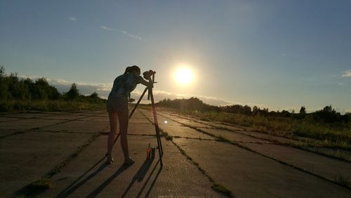 Full length of woman photographing with tripod on footpath against sky during sunset