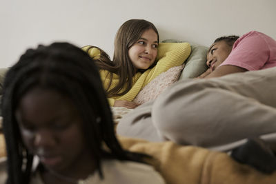 Teenage girls talking while lying in bed by female friend sitting in front at home