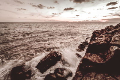 Scenic view of sea waves splashing on rocks against sky during sunset