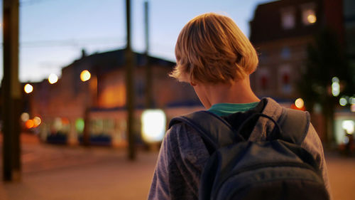 Rear view of boy standing with backpack in city at dusk