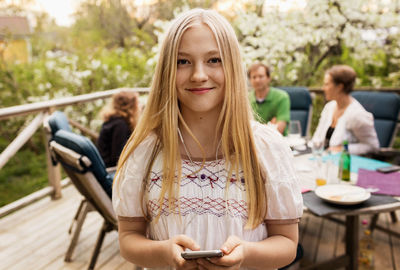 Portrait of teenage girl holding smart phone at yard with family sitting in background